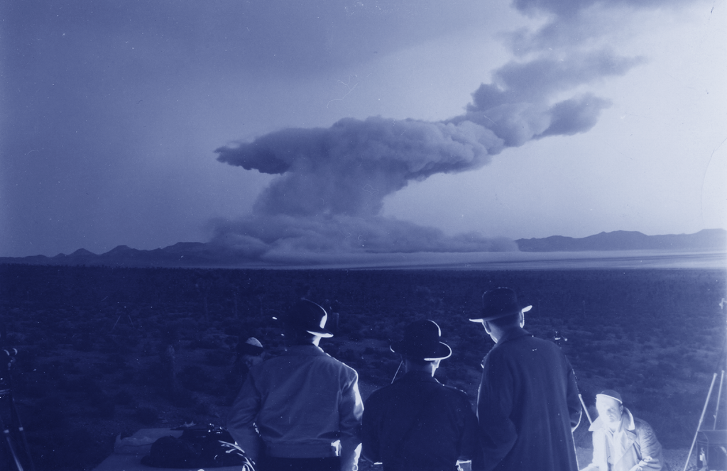 This is an image from the Trinity nuclear test. Three men in hats watch the mushroom cloud from afar.